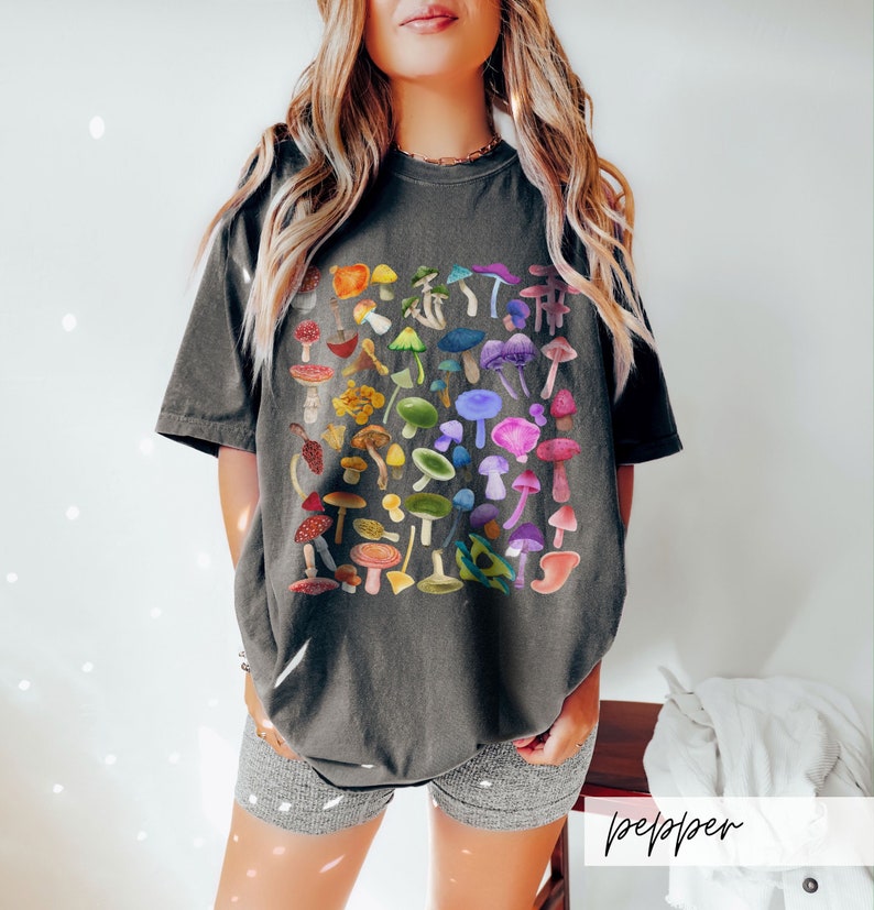 K&ouml;nnte beinhalten: Ein dunkelgraues T-Shirt mit einem bunten Pilzdruck. Die Pilze sind in einem quadratischen Muster angeordnet und in verschiedenen Schattierungen von Rot, Orange, Gelb, Gr&uuml;n, Blau, Lila und Rosa.