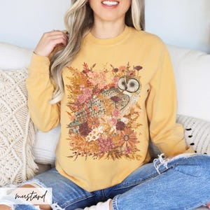 Pode incluir: Uma camisa de moletom amarela mostarda com uma estampa gr&aacute;fica de uma coruja cercada por flores de outono.