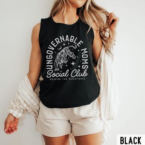 Puede incluir: Camiseta sin mangas negra con un gr&aacute;fico blanco que presenta un tigre, una luna creciente, estrellas y el texto "YOUNGGOVERNABLE MOMS Social Club RAISING THE RESISTANCE". La palabra "BLACK" est&aacute; en la esquina inferior derecha.