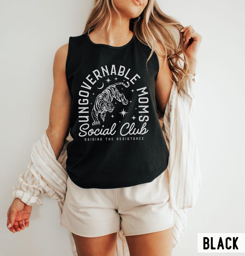 Puede incluir: Camiseta sin mangas negra con un gr&aacute;fico blanco que presenta un tigre, una luna creciente, estrellas y el texto "YOUNGGOVERNABLE MOMS Social Club RAISING THE RESISTANCE". La palabra "BLACK" est&aacute; en la esquina inferior derecha.