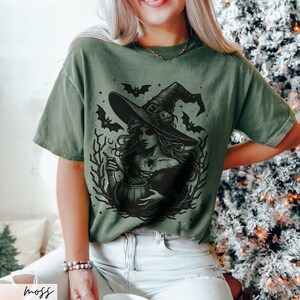 May include: A green t-shirt with a black and white illustration of a witch stirring a cauldron. The witch is wearing a pointed hat and has long, flowing hair. Bats and stars are in the background.