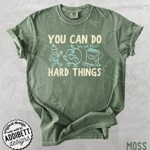 K&ouml;nnte beinhalten: Moosgr&uuml;nes T-Shirt mit dem wei&szlig;en Text "YOU CAN DO HARD THINGS". Das Design umfasst Cartoon-Illustrationen eines Bleistifts, eines Apfels und eines Buches. Das Shirt hat auch den Text "you got this!" &uuml;ber dem Wort "HARD".