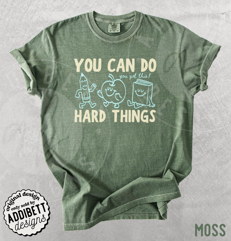 K&ouml;nnte beinhalten: Moosgr&uuml;nes T-Shirt mit dem wei&szlig;en Text "YOU CAN DO HARD THINGS". Das Design umfasst Cartoon-Illustrationen eines Bleistifts, eines Apfels und eines Buches. Das Shirt hat auch den Text "you got this!" &uuml;ber dem Wort "HARD".
