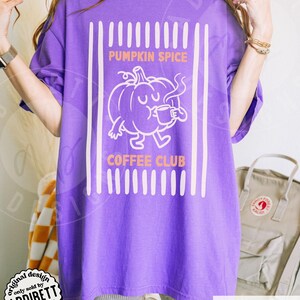 May include: A violet short-sleeved t-shirt featuring a white graphic of a pumpkin character drinking from a mug. The text reads PUMPKIN SPICE COFFEE CLUB in orange letters, framed by vertical white stripes on a solid violet background fabric.
