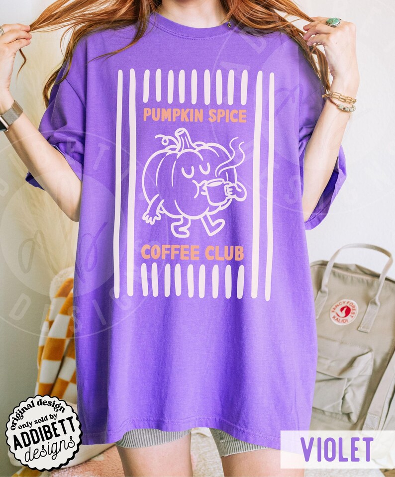 May include: A violet short-sleeved t-shirt featuring a white graphic of a pumpkin character drinking from a mug. The text reads PUMPKIN SPICE COFFEE CLUB in orange letters, framed by vertical white stripes on a solid violet background fabric.