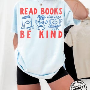 May include: A light blue t-shirt featuring the red text READ BOOKS and BE KIND. Between the text are three blue cartoon characters: a smiling book, a flower in a pot, and an open book, with the blue text stay weird written above them.