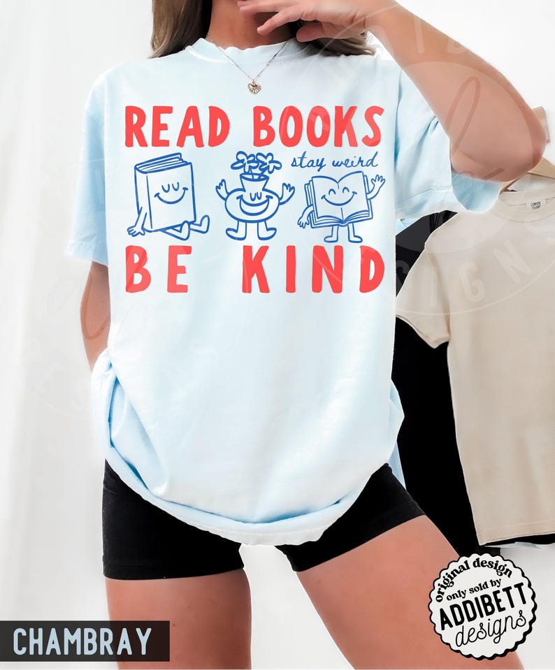 May include: A light blue t-shirt featuring the red text READ BOOKS and BE KIND. Between the text are three blue cartoon characters: a smiling book, a flower in a pot, and an open book, with the blue text stay weird written above them.