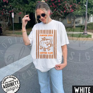 May include: A white short-sleeve t-shirt featuring a brown graphic print of a cartoon pumpkin holding a coffee mug. The text PUMPKIN SPICE COFFEE CLUB is centred above and below the pumpkin, framed by vertical brown stripes on both sides.