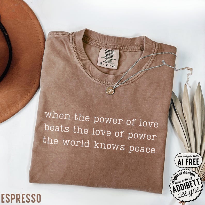 May include: A brown short-sleeved t-shirt with the white printed text: when the power of love beats the love of power the world knows peace. The shirt features a crew neckline and a Comfort Colors brand label on the inside collar.