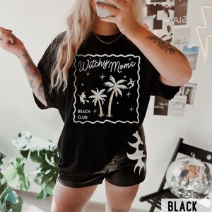 K&ouml;nnte beinhalten: Schwarzes T-Shirt mit einem wei&szlig;en Aufdruck &bdquo;Witchy Moms Beach Club&ldquo;. Die Grafik zeigt Palmen, Sterne und Silhouetten von Hexen. Das Wort &bdquo;BLACK&ldquo; ist in einem wei&szlig;en Rechteck am unteren Bildrand aufgedruckt.