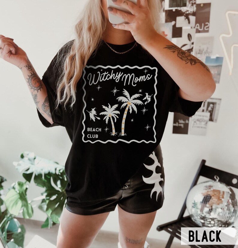 K&ouml;nnte beinhalten: Schwarzes T-Shirt mit einem wei&szlig;en Aufdruck &bdquo;Witchy Moms Beach Club&ldquo;. Die Grafik zeigt Palmen, Sterne und Silhouetten von Hexen. Das Wort &bdquo;BLACK&ldquo; ist in einem wei&szlig;en Rechteck am unteren Bildrand aufgedruckt.