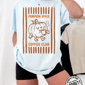 May include: A light blue t-shirt featuring a brown graphic print of a cartoon pumpkin holding a coffee mug. The text reads PUMPKIN SPICE COFFEE CLUB. The design is framed by vertical brown stripes. The shirt is paired with black shorts.