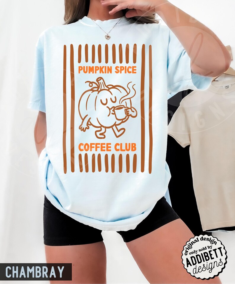 May include: A light blue t-shirt featuring a brown graphic print of a cartoon pumpkin holding a coffee mug. The text reads PUMPKIN SPICE COFFEE CLUB. The design is framed by vertical brown stripes. The shirt is paired with black shorts.