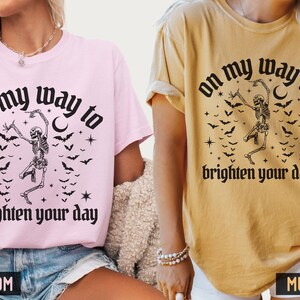 May include: Two t-shirts, one pink and one mustard, each with a black graphic. The graphic features a dancing skeleton, bats, stars, and the text "on my way to brighten your day."