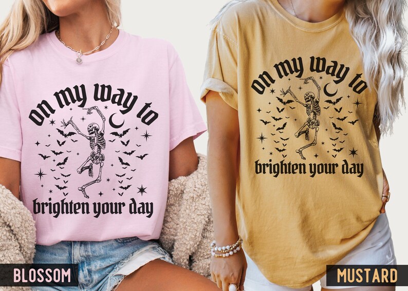 May include: Two t-shirts, one pink and one mustard, each with a black graphic. The graphic features a dancing skeleton, bats, stars, and the text "on my way to brighten your day."
