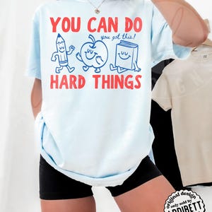 K&ouml;nnte beinhalten: Hellblaues T-Shirt mit rotem Text "YOU CAN DO HARD THINGS" und dem blauen Satz "you got this!". Das Shirt zeigt Cartoon-Illustrationen eines Bleistifts, Apfels und Buches. Original-Design, verkauft von Addibett Designs.