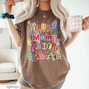 May include: Espresso-colored t-shirt with the text "Weird Moms Build Character" in a colorful, collage-style design. The shirt features various graphics, including flowers, lightning bolts, and a checkered heart. A mug with text is held in the hand.