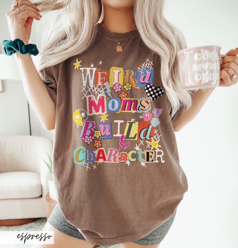 May include: Espresso-colored t-shirt with the text "Weird Moms Build Character" in a colorful, collage-style design. The shirt features various graphics, including flowers, lightning bolts, and a checkered heart. A mug with text is held in the hand.