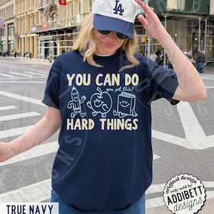 K&ouml;nnte beinhalten: Marineblaues T-Shirt mit dem Text "YOU CAN DO HARD THINGS" und Cartoon-Illustrationen eines Bleistifts, Apfels und Buches. Das Shirt hat auch den Text "you got this!" &uuml;ber den Illustrationen. Das Shirt ist von ADDIBETT designs.