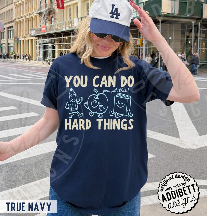 K&ouml;nnte beinhalten: Marineblaues T-Shirt mit dem Text "YOU CAN DO HARD THINGS" und Cartoon-Illustrationen eines Bleistifts, Apfels und Buches. Das Shirt hat auch den Text "you got this!" &uuml;ber den Illustrationen. Das Shirt ist von ADDIBETT designs.