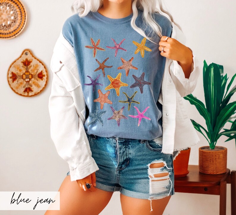 May include: A blue jean colored t-shirt featuring a collection of colorful starfish designs. The starfish are in various colors, including orange, pink, yellow, and purple. The text "blue jean" is at the bottom of the image.