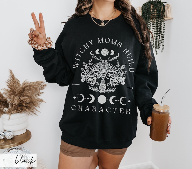 K&ouml;nnte beinhalten: Schwarzes Sweatshirt mit dem wei&szlig;en Text "Witchy Moms Build Character". Das Design umfasst eine Motte, Halbmonde und ein Pentagramm. Das Sweatshirt hat einen Rundhalsausschnitt.
