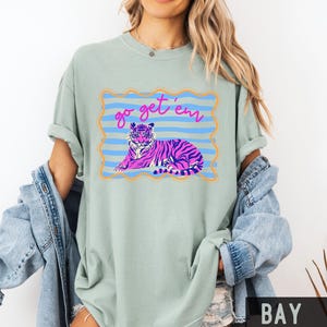 Pu&ograve; includere: T-shirt verde chiaro con un disegno di tigre rosa e viola all'interno di una cornice blu e arancione. Il testo "go get 'em" &egrave; sopra la tigre. La maglietta ha maniche arrotolate e girocollo.