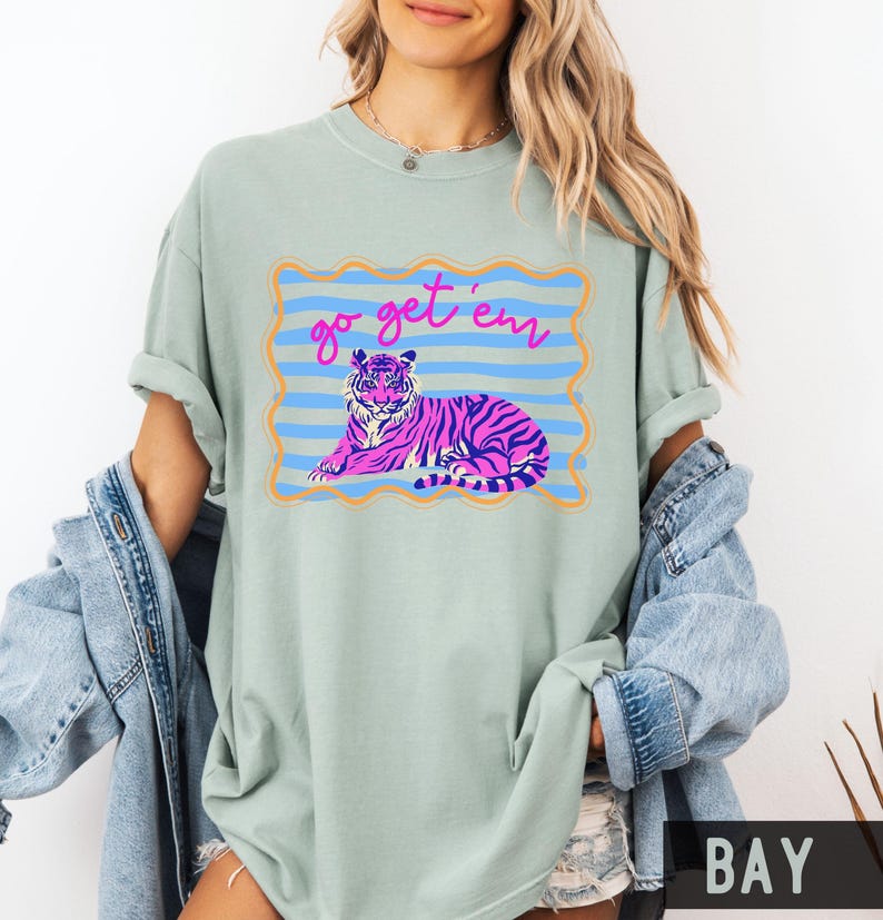 Pu&ograve; includere: T-shirt verde chiaro con un disegno di tigre rosa e viola all'interno di una cornice blu e arancione. Il testo "go get 'em" &egrave; sopra la tigre. La maglietta ha maniche arrotolate e girocollo.