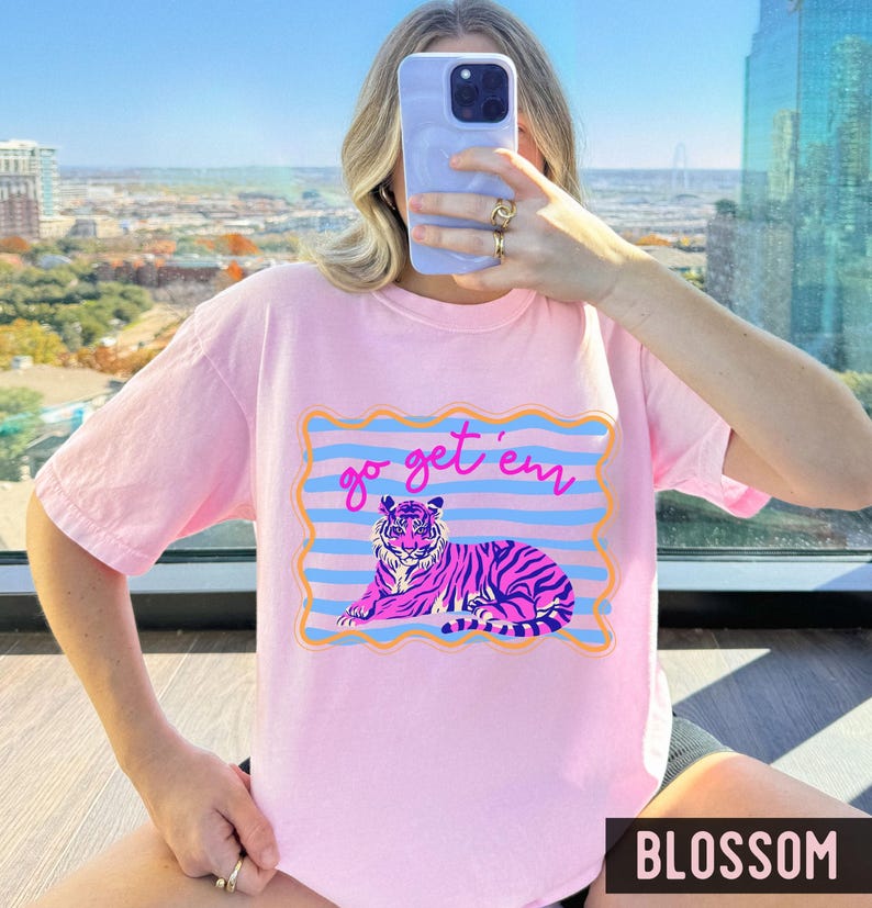 Pu&ograve; includere: T-shirt rosa chiaro con grafica di tigre e la scritta "go get 'em". La tigre &egrave; raffigurata in rosa e viola, con dettagli blu e arancioni. La parola "BLOSSOM" &egrave; stampata in basso.