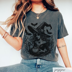 May include: A dark gray t-shirt with a black and white illustration of a witch stirring a cauldron. The witch is wearing a pointed hat and has long flowing hair. There are bats and stars in the background.