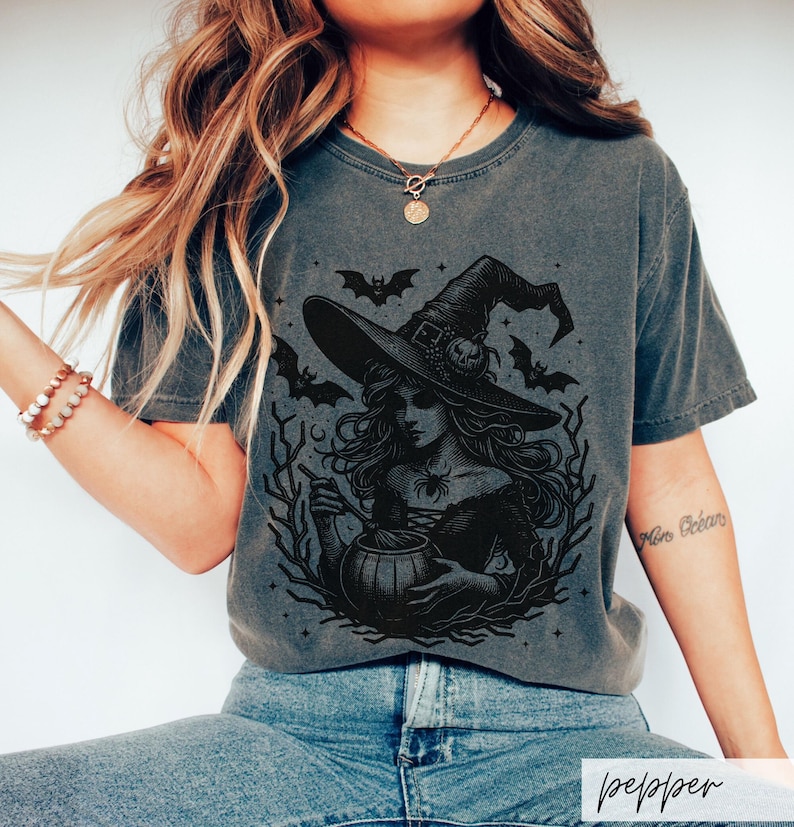 May include: A dark gray t-shirt with a black and white illustration of a witch stirring a cauldron. The witch is wearing a pointed hat and has long flowing hair. There are bats and stars in the background.