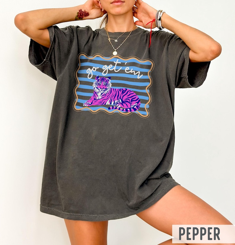 Pu&ograve; includere: T-shirt grigio scuro con una grafica di una tigre viola e rosa, strisce blu e la scritta "go get 'em". Il disegno &egrave; incorniciato da un bordo ondulato. La maglietta ha una vestibilit&agrave; ampia.