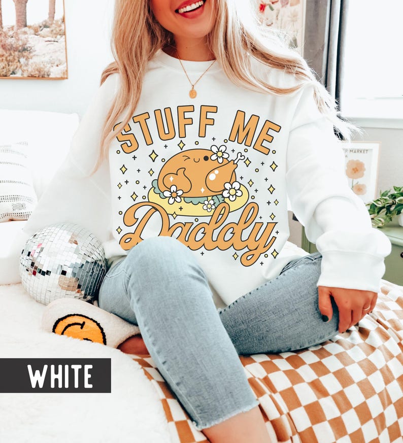 Pu&ograve; includere: Felpa bianca con grafica retr&ograve; di un tacchino arrosto su un piatto giallo, circondato da fiori e stelle. Il testo recita "Stuff Me Daddy" in arancione. Uno stile casual e confortevole.