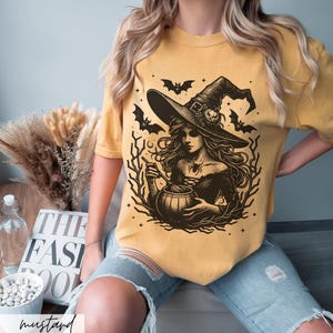 May include: A mustard yellow t-shirt with a black and white illustration of a witch stirring a cauldron. The witch is wearing a pointed hat and has long, flowing hair. Bats and stars are in the background.
