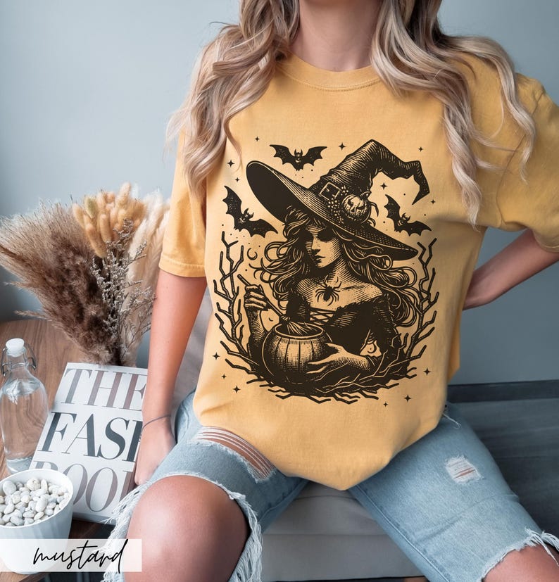 May include: A mustard yellow t-shirt with a black and white illustration of a witch stirring a cauldron. The witch is wearing a pointed hat and has long, flowing hair. Bats and stars are in the background.