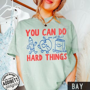 K&ouml;nnte beinhalten: Hellgr&uuml;nes T-Shirt mit dem roten Schriftzug "YOU CAN DO HARD THINGS". Das Design zeigt Cartoon-Zeichnungen eines Bleistifts, eines Apfels und eines Buches. Der Satz "you got this!" ist ebenfalls enthalten. Das T-Shirt hat einen Rundhalsausschnitt.
