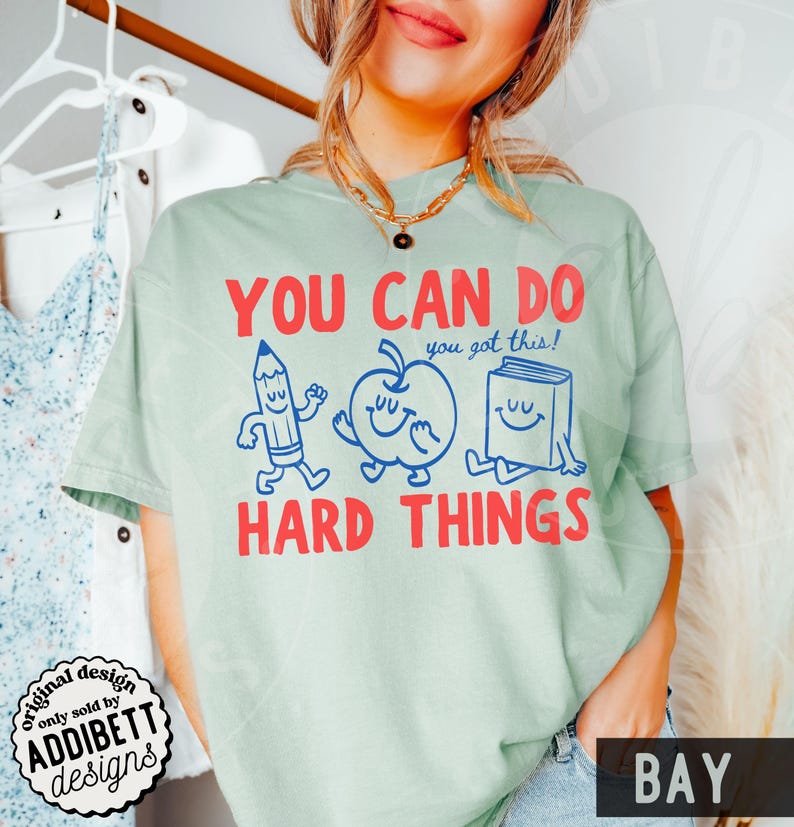 K&ouml;nnte beinhalten: Hellgr&uuml;nes T-Shirt mit dem roten Schriftzug "YOU CAN DO HARD THINGS". Das Design zeigt Cartoon-Zeichnungen eines Bleistifts, eines Apfels und eines Buches. Der Satz "you got this!" ist ebenfalls enthalten. Das T-Shirt hat einen Rundhalsausschnitt.