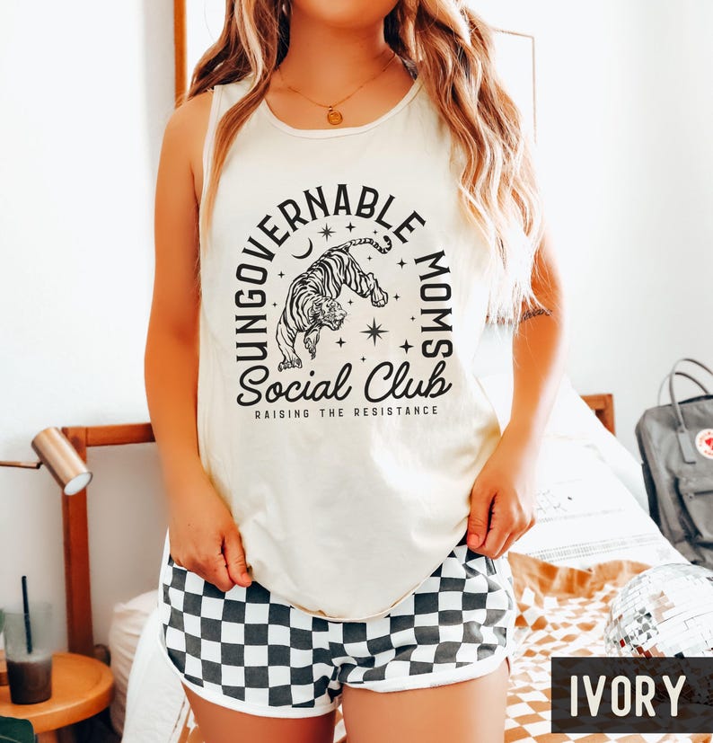 Puede incluir: Camiseta sin mangas marfil con un gr&aacute;fico de tigre en blanco y negro y el texto "UNGOVERNABLE MOMS SOCIAL CLUB RAISING THE RESISTANCE". La modelo tambi&eacute;n lleva shorts a cuadros blancos y negros.
