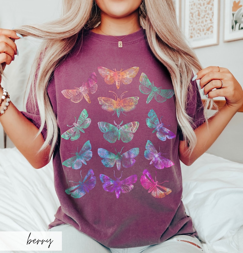 May include: A purple t-shirt featuring a colourful butterfly print. The butterflies are arranged in a symmetrical pattern and are all different colours and sizes.
