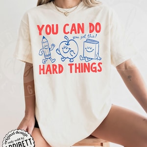K&ouml;nnte beinhalten: Cremefarbenes T-Shirt mit rotem Text "YOU CAN DO HARD THINGS". Das Design zeigt einen blauen Bleistift, Apfel und Buch mit l&auml;chelnden Gesichtern und dem Satz "you got this!". Das T-Shirt stammt von "ADDIBETT designs".