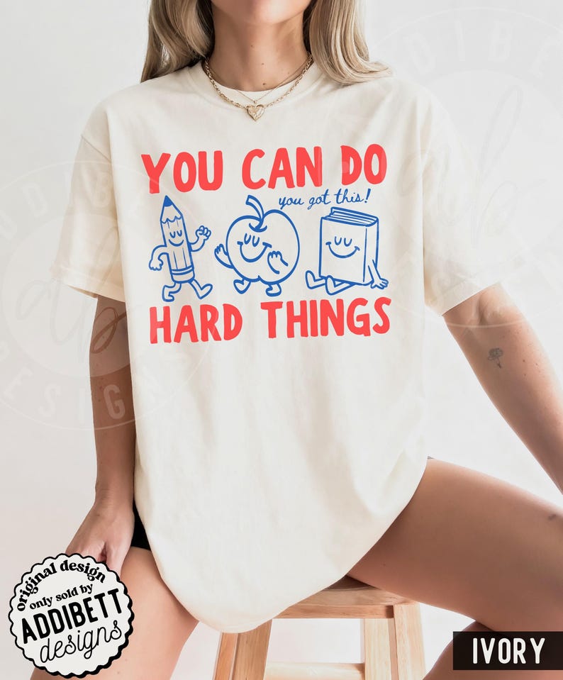 K&ouml;nnte beinhalten: Cremefarbenes T-Shirt mit rotem Text "YOU CAN DO HARD THINGS". Das Design zeigt einen blauen Bleistift, Apfel und Buch mit l&auml;chelnden Gesichtern und dem Satz "you got this!". Das T-Shirt stammt von "ADDIBETT designs".