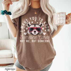 以下が含まれることがあります： 茶色のTシャツに、サングラスをかけたアライグマと「誰かが気にかけていることを知ってほしい。私じゃない、誰か。」という文字がプリントされています。