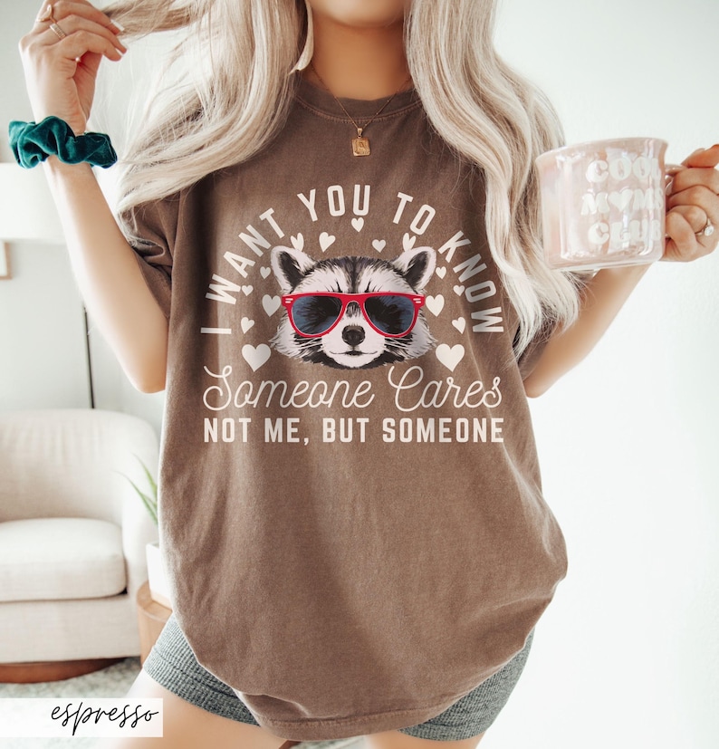 以下が含まれることがあります： 茶色のTシャツに、サングラスをかけたアライグマと「誰かが気にかけていることを知ってほしい。私じゃない、誰か。」という文字がプリントされています。