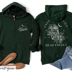 May include: Forest green zip-up hoodie with a white graphic of an open book and crescent moon on the front. The back features a stack of books, flowers, and the text "FREE SOCIETIES READ FREELY". The hoodie has a hood and front pockets.