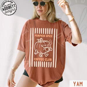 May include: A rust-coloured short-sleeve t-shirt featuring a white graphic of a cartoon pumpkin drinking from a mug. The text reads PUMPKIN SPICE COFFEE CLUB, framed by vertical white stripes. The design is centred on the front of the garment.