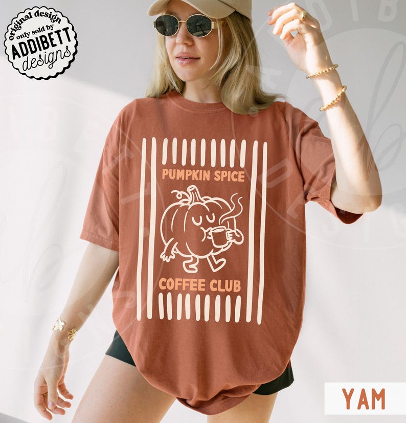 May include: A rust-coloured short-sleeve t-shirt featuring a white graphic of a cartoon pumpkin drinking from a mug. The text reads PUMPKIN SPICE COFFEE CLUB, framed by vertical white stripes. The design is centred on the front of the garment.