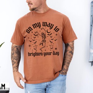 May include: A terracotta-coloured t-shirt with a black graphic of a dancing skeleton, bats, and the words "on my way to brighten your day." The shirt is being worn by a person.