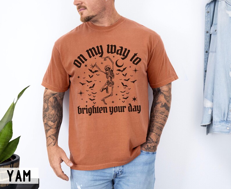 May include: A terracotta-coloured t-shirt with a black graphic of a dancing skeleton, bats, and the words "on my way to brighten your day." The shirt is being worn by a person.