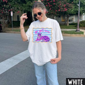 Pu&ograve; includere: T-shirt bianco con grafica di una tigre viola, dettagli blu e rosa, e la scritta "go get 'em". Il disegno &egrave; incorniciato da un bordo blu e giallo. La maglietta &egrave; in stile casual e unisex.