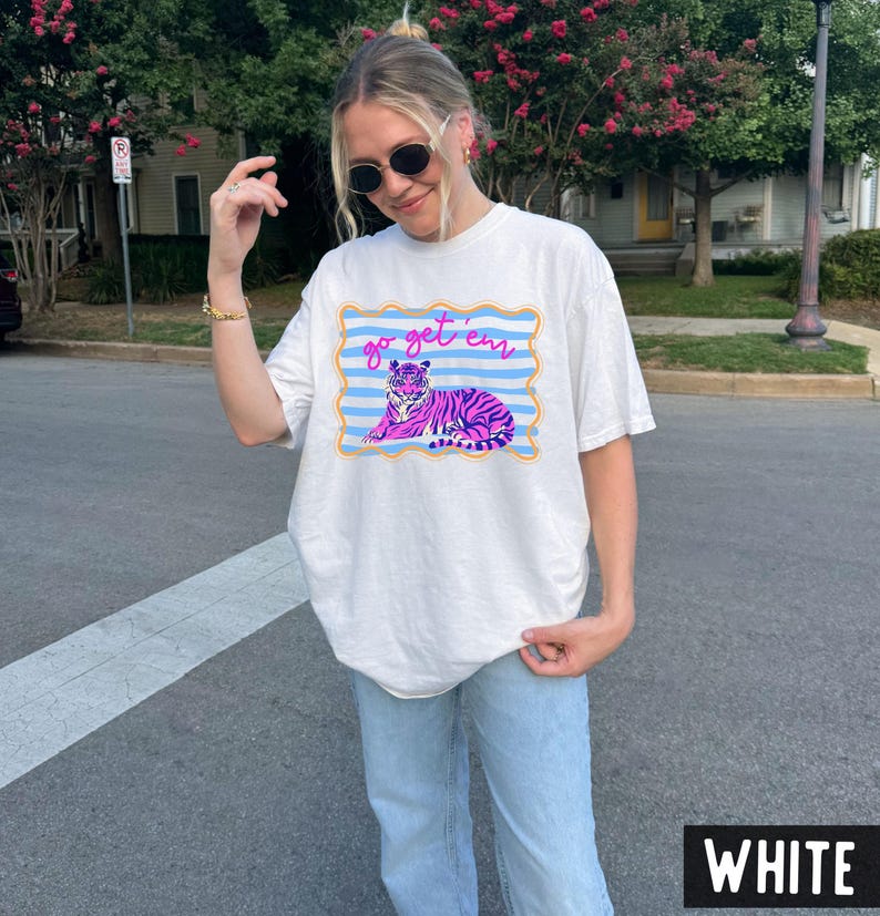 Pu&ograve; includere: T-shirt bianco con grafica di una tigre viola, dettagli blu e rosa, e la scritta "go get 'em". Il disegno &egrave; incorniciato da un bordo blu e giallo. La maglietta &egrave; in stile casual e unisex.